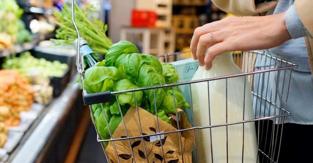 A person shops in a grocery store with a basket containing fresh greens, milk, and herbs.
